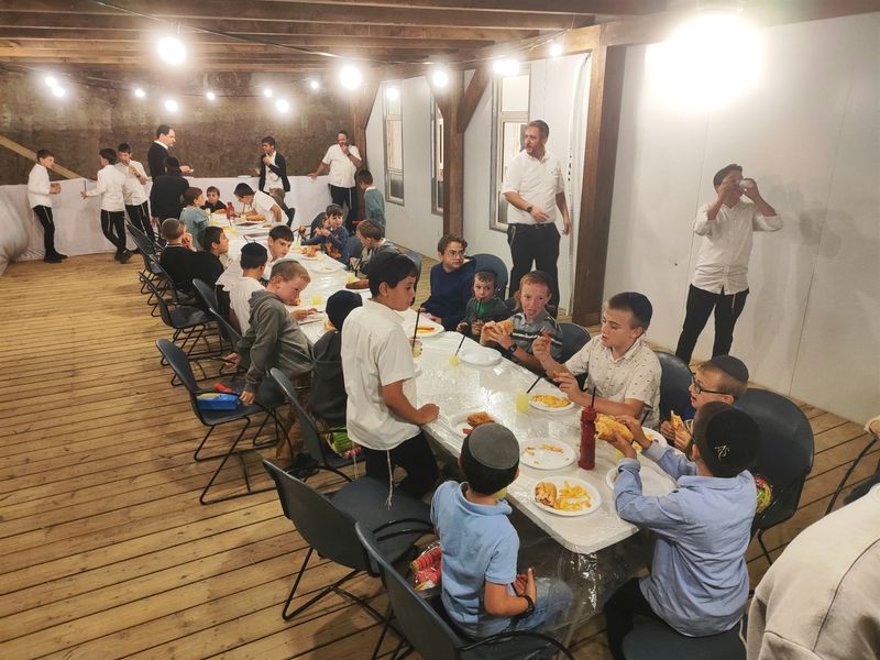 ילדים יושבים סביב שולחן ארוך בפרגולה של בית הכנסת, תחת שרשרת נורות