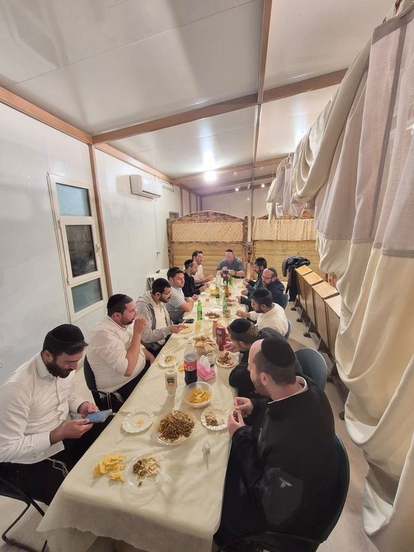 אנשי הקהילה יושבים לסעודה סביב שולחן ארוך בתוך בית הכנסת